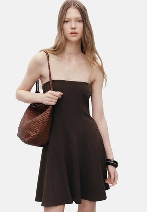 Brown strapless dress with a fitted bodice and flared skirt. Model holds a woven brown bag. Smooth fabric with clean lines and minimalistic design.