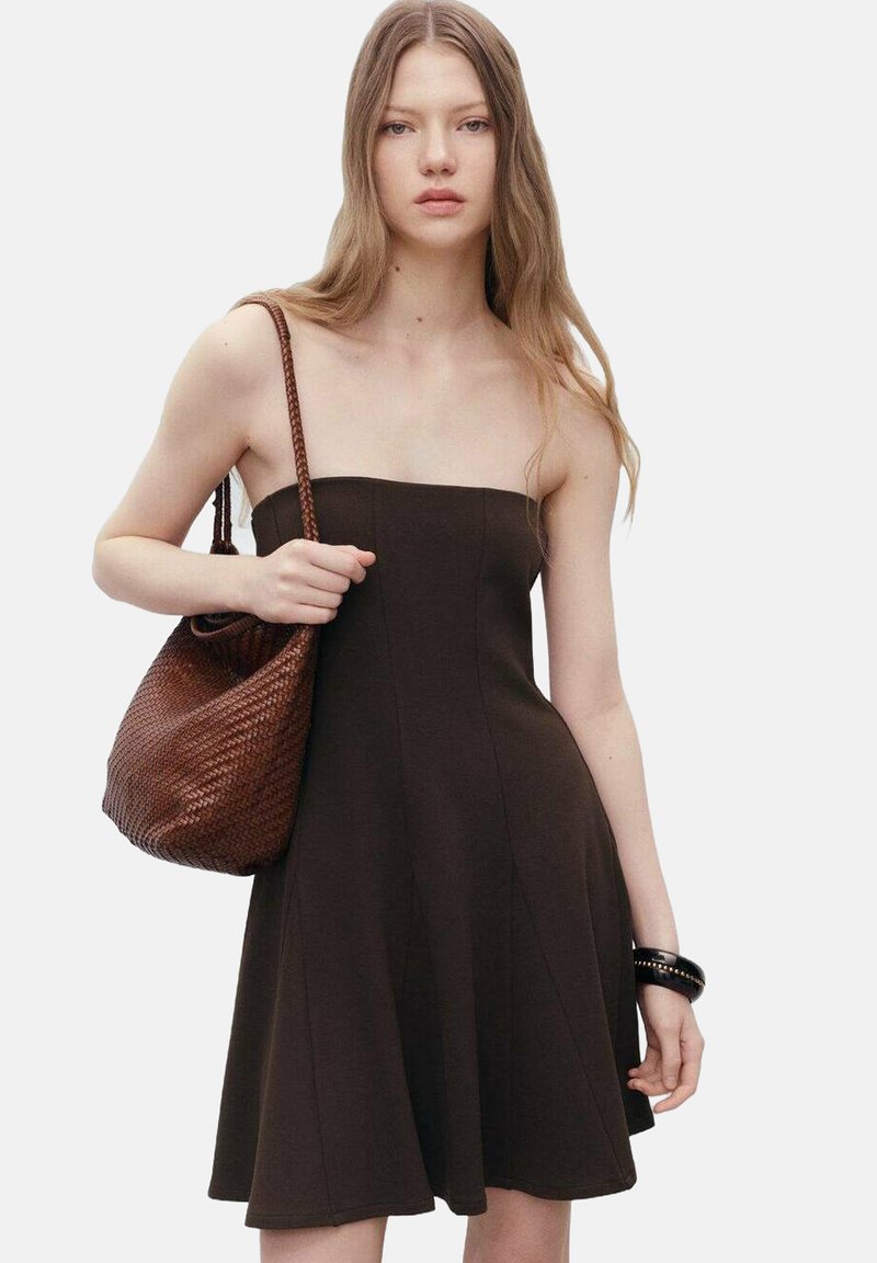 Braunes trägerloses Kleid mit tailliertem Oberteil und ausgestelltem Rock. Das Model hält eine geflochtene braune Tasche. Glattes Material mit klaren Linien und minimalistischem Design.