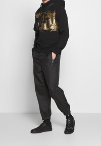 Černá mikina s kapucí se zlatým grafickým motivem, spojená s černými lehkými tepláky s elastickými manžetami a bočními zipy, doplněná černými teniskami.