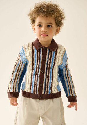 Junges Kind trägt eine braun-creme-blaue, gestreifte Strickjacke mit Knöpfen und beige Hosen, steht vor einem hellen Hintergrund.