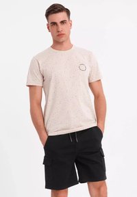 Lichtbeige t-shirt met zwarte spikkels en een klein embleem op de borst, gecombineerd met zwarte cargoshorts. Eenvoudig ontwerp en relaxte pasvorm.
