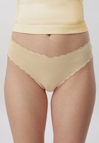 Femme portant des sous-vêtements beige sans couture avec des bords festonnés et un haut assorti beige, montrée de la taille jusqu'aux cuisses supérieures.