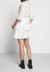 Valkoinen kerroksittain leikattu mekko, jossa on silmäreikäkangas, lyhyet röyhelöhihares ja istuva vyötärö. Yhdistetty mustien nilkkureiden kanssa.