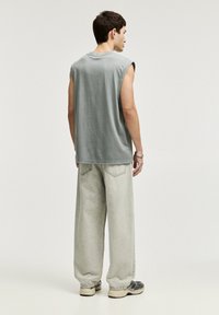 Mouwloze grijze katoenen shirt met ruwe randen, gecombineerd met losse beige denim jeans. De outfit heeft een relaxte pasvorm en een casual stijl.