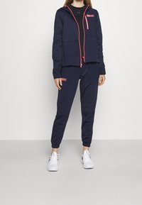 Marineblaue Kapuzenjacke mit pinkfarbenen Reißverschlussakzenten, kombiniert mit passenden Jogginghosen. Verfügt über eine Fronttasche und gerippte Bündchen. Weiße Sportschuhe.