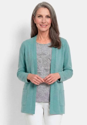 Lachende vrouw met grijs haar die een licht turquoise vest draagt over een grijs topje met patroon en een witte broek, staand tegen een effen achtergrond.