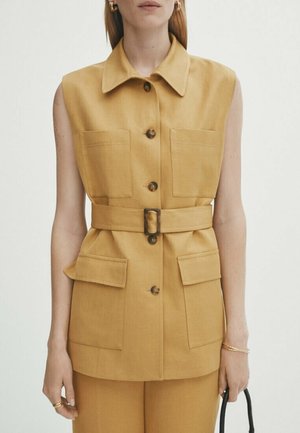 Tan sleeveless vest with a collar, button front, two chest pockets, belted waist, and a textured fabric finish. Accented with large buttons.