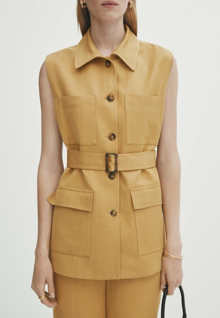 Tan ärmlös väst med krage, knappstängning fram, två bröstfickor, belagd midja och en texturerad tygfinish. Accenterad med stora knappar.