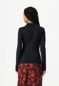 Svart langermet polo skjorte med krage, tett passform. Den nedre kanten faller over en rød og svart skjørt med blomsterdesign. Glatt tekstur.