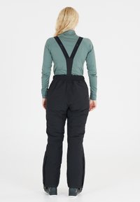 Zwarte skibroeken met verstelbare bretels, elastische tailleband en ritsopeningen aan de zijkant, gecombineerd met een groene lange mouw top.