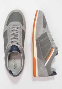 Grå sportskor i nubuck och mesh-material, med orange detaljer, snörningsdesign och en gummisula med en kontrasterande rand.