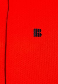 Röd textil med en strukturerad yta och en liten, broderad svart logotyp som visar ett stiliserat "B" och vertikala linjer.