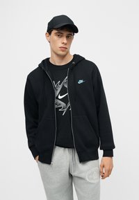 Νέος άνδρας που φοράει μαύρο καπέλο Nike, μαύρη φούτερ ζακέτα με φερμουάρ και μπλε λογότυπο, μαύρο μπλουζάκι με σχέδιο και ανοιχτό γκρι φόρμα με λευκό έμβλημα.