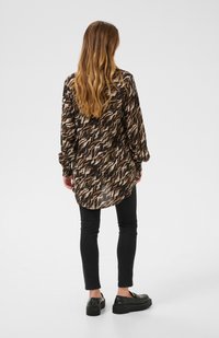 Camicia a maniche lunghe con un motivo ondulato marrone e crema, realizzata in tessuto leggero, con orlo curvo e polsini arricciati. Abbinata a pantaloni neri.