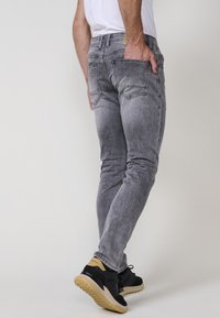 Šedé slim-fit džíny vyrobené z denimu s vytíraným povrchem, vybavené pěti kapsami a červenými stehy, v kombinaci s černými sportovními botami.
