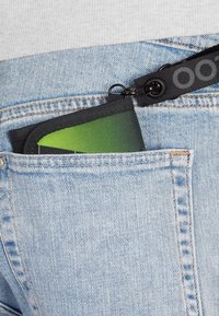 Ein grüner und schwarzer Beutel, der teilweise in einer Jeanstasche sichtbar ist, aus einem strukturierten Material mit einem schwarzen Riemen und einer Hardware-Befestigung.