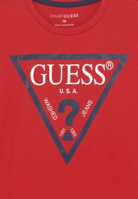 Röd bomull t-shirt med en triangulär grafisk design i marin. Den centrala logotypen har texten "GUESS," med "WASHED" och "JEANS" bredvid ett frågetecken.