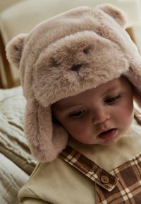 Minkštas smėlio spalvos meškinas kepurė su purus tekstūra, su mažomis ausytėmis ir veido piešiniu. Derinama su blankiai geltonu rūbu ir languotais puskombinezoniais.