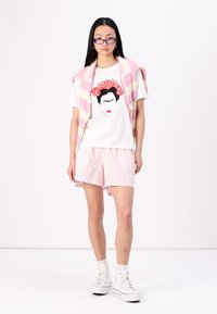 Hvit T-skjorte med Frida Kahlo-grafikk, paret med rosa stripete shorts og en rosa stripete genser drapert over skuldrene; iført hvite sneakers.