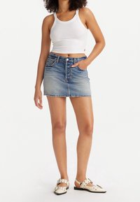 Weißes, geripptes Crop-Top, gepaart mit einem hellblauen Jeans-Minirock mit fransigem Saum, der aufgesetzte Taschen und einen Knopf am Bund hat.