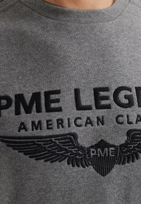 Sudadera gris hecha de tela texturizada, con un logotipo bordado negro que presenta un diseño de alas y el texto "PME LEGEND AMERICAN CLASSIC".