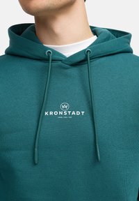 Sudadera de color verde azulado hecha de un tejido suave, con un logo frontal "KRONSTADT" en blanco. Los cordones y un bolsillo tipo canguro completan el diseño.
