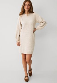 Kvinde står foran en ensfarvet baggrund iført en beige langærmet strikkjole og brune flade sko med spænde detaljer.