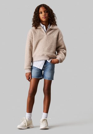 Pullover beige soffice con colletto, indossato sopra una camicia a righe bianche, abbinato a pantaloni corti di denim blu e scarpe da ginnastica bianche con calzini al polpaccio.