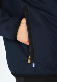 Marineblå softshell jakke med en lynlåslomme, der har en lædertrækning og et accent med det svenske flag. Materialet er glat og let.