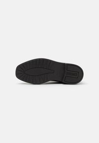 Semelle de chaussure en caoutchouc noir avec une surface texturée, comportant un motif de bande de roulement pour l'adhérence. Logo embossé sur le côté, zone de talon rectangulaire.