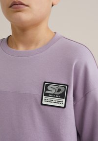Violeta sporta krekls ar teksturētu apakšējo daļu un melnbaltu "Salty Dog" logo uzšuvumu uz krūtīm. Ribināta apkakle un maiga auduma kompozīcija.