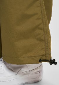 Bas de pantalon kaki avec cordon noir ajustable au-dessus d'une chaussure Nike Air blanche sur fond gris clair.