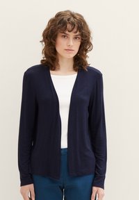Donkerblauwe lichte cardigan van zachte stof, met een open voorkant, lange mouwen, gedragen over een witte top met een ronde halslijn.