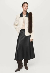 Giacca bianca con colletto nero, indossata sopra un top beige. Gonna midi in pelle nera, abbinata a stivali alti fino alla coscia marroni. Accentatura in pelliccia marrone applicata.