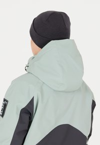 Vattentålig jacka i mintgrön med grå detaljer. Har en huva, minimal logotyp på ärmen och en slät yta. Mörk stickad mössa synlig.