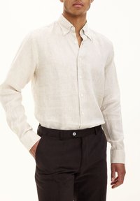Camicia in lino beige chiaro con colletto button-down, maniche lunghe e taglio morbido, abbinata a pantaloni scuri con chiusura a bottoni.