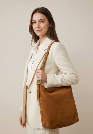 Vrouw in beige pak met een grote bruine suède schoudertas met metalen studs tegen een effen achtergrond.