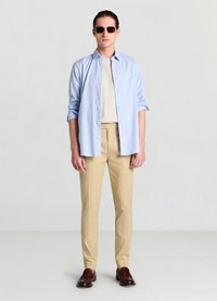 Hellblau-weiß gestreiftes Hemd mit Knöpfen, beigefarbene Hose und braune Loafers. Das Hemd wird offen über einem neutralen Oberteil getragen, dazu Sonnenbrille.