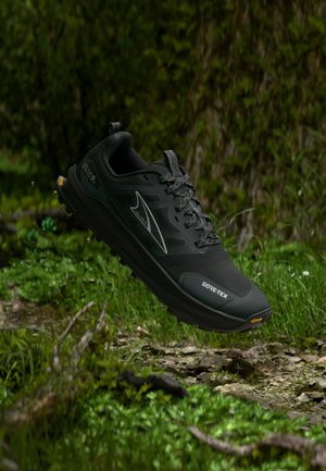 Czarny but do biegania po szlakach Gore-Tex zawieszony nad trawiastym, kamienistym leśnym podłożem, wokół podeszwy unoszą się cząsteczki ziemi w ruchu.