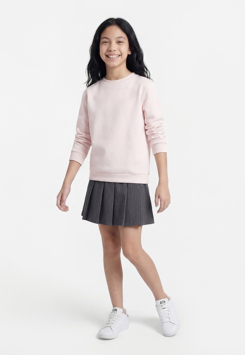 Pige med langt sort hår, der smiler, iført lyserød sweatshirt, mørkegråt plisseret skørt og hvide sneakers, stående foran en hvid baggrund.