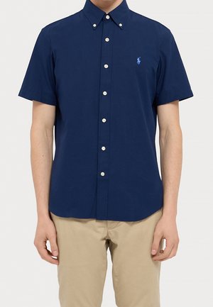 Mężczyzna ubrany w granatową koszulę z krótkim rękawem zapinaną na guziki z małym haftowanym logo na piersi oraz beżowe spodnie.