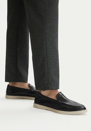 Schwarze Leder-Slipper mit beigefarbenen Sohlen, getragen zu grauen, gerade geschnittenen Hosen, vor weißem Hintergrund abgebildet.