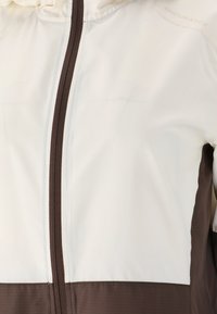 Lichte jas in off-white en bruin, met een ritssluiting over de gehele lengte en contrasterende kleurblokken. Soepele textuur met subtiele branding.