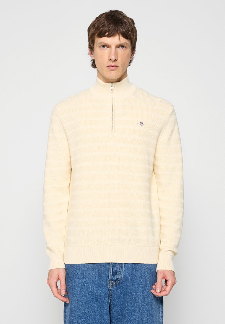 Homme aux cheveux bruns courts portant un pull crème à zip quart et un jean bleu ample, se tenant devant un fond clair uni.