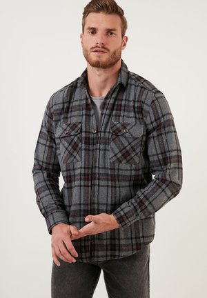 Camisa de franela gris con un patrón de cuadros rojo y negro, dos bolsillos en el pecho y detalles de botones. Cuello clásico, mangas largas y textura suave.