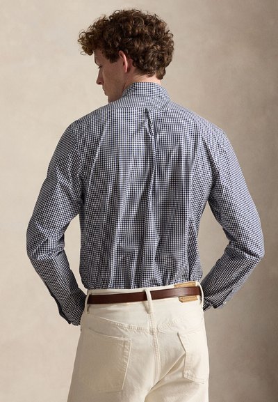 Homme aux cheveux bouclés vu de dos, portant une chemise à carreaux bleue à manches longues rentrée dans un pantalon écru avec une ceinture marron.