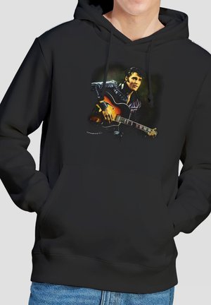 Person trägt schwarzen Hoodie mit Grafik eines Mannes, der akustische Gitarre spielt, Hände in den Taschen des Hoodies, kombiniert mit blauen Jeans.