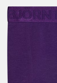 Lila midjeband med präglad "BJÖRN"-logotyp. Materialet är slätt stickat tyg som visar en konsekvent struktur och färg genom hela.