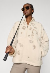 Πουλόβερ φλις ζακέτα σε κρεμ απόχρωση με φερμουάρ του ενός τετάρτου. Διαθέτει πασλιέ κεντήματα σε μπεζ και μπροστινή τσέπη. Το μοντέλο κρατάει ένα μαύρο αντικείμενο.