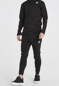 Schwarzer sportlicher Trainingsanzug aus glattem, dehnbarem Stoff. Verfügt über eine Jacke mit vollem Reißverschluss und Kragen sowie enganliegende Hosen mit elastischen Bündchen.